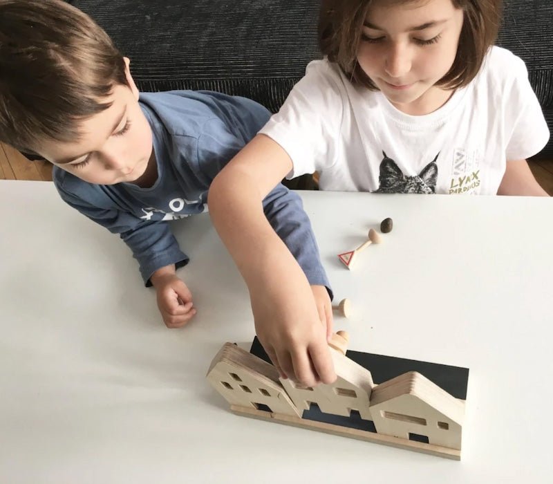 Dos niños jugando con unas casitas de madera, sobre una mesa blanca.